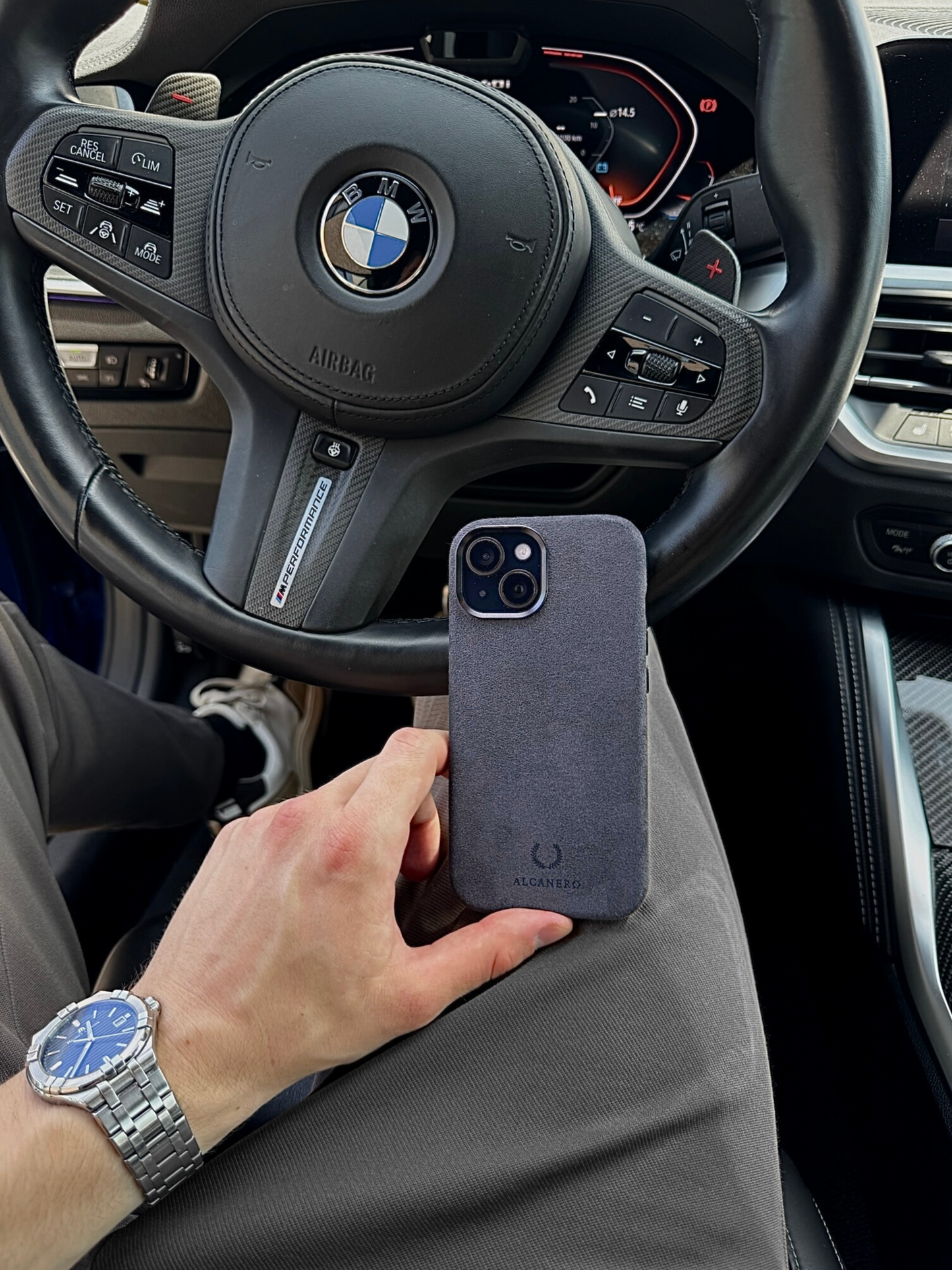 Black Alcantara iPhone 15 case by Alcanero held in front of a BMW M Performance steering wheel – stylish accessory for business and lifestyle.